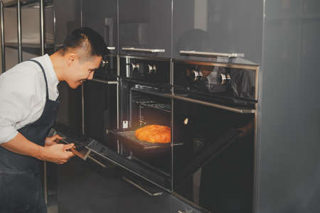 Young man baker opens a bread oven in a modern kitchen at home : Happy and smiling Asian man...