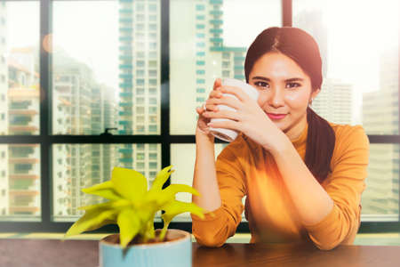 Portrait of a young and beautiful business woman sitting with a glass of good mood while sitting and relaxing. Relaxing in the office by the window in a relaxed atmosphere, looking at the camera.の写真素材