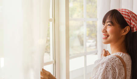 Good morning new day : Asian cute girl fairy opens the window curtain greets the morning sun in the living room looks outside smiling has a good attitude and health.の写真素材