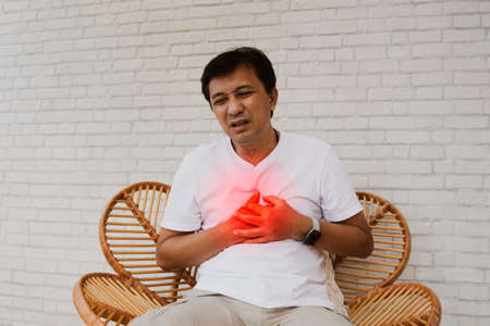 Senior portrait Asian middle-aged man with sudden cardiac disease showing pain leaky heart muscle tired and suffocating sat wooden chair in agony : Myocardial infarction (Heart Attの写真素材
