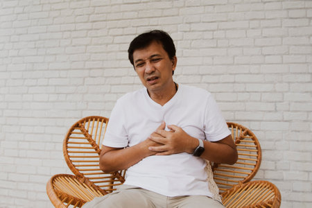 Middle aged asian senior man with heart disease showing pain, myocardial leak, weakness, fatigue and shortness of breath, sits with his hands over his chest in agony.の写真素材