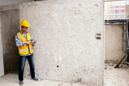 Asian male architect wearing helmet with laptop inspects construction site to renovate the bathroom according to the blueprint design : Engineer worker pay attention to building woの写真素材