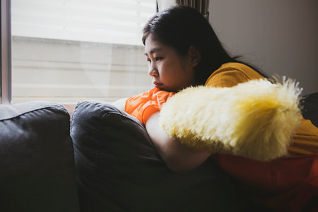 Teenage girl with duster sat idle on the sofa looking at the window bored from the boring chores resting because she was too lazy to do housework and was alone in the house.の写真素材