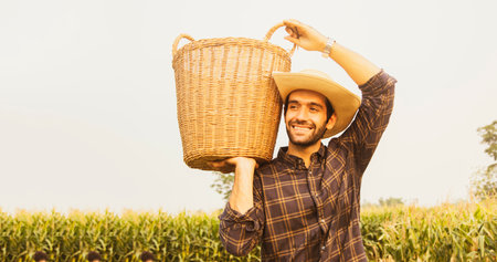 Joyful male farmer looked at the corn farm with smile and proudly carrying basket on his shoulder to work to collect some of the crops he had planted in the corn field.の写真素材