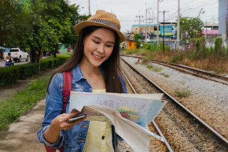 Beautiful asian woman backpack stands excitedly looking interesting tourist map while waiting for the train at the suburban station to pick up her departure and special train trip.の写真素材