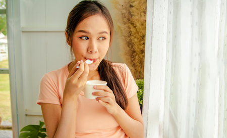 Portrait cute long haired asian woman eating sweet milk ice cream from cup standing next to a sunlit window at home : Smiling girl looking at the camera eating ice cream with a spoの写真素材