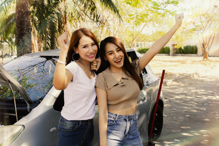 Portrait two carefree asian women parked on the sidewalk enjoying broken car and flat tire as they travel, showing smiles and comfort waiting for help from mechanic.の写真素材