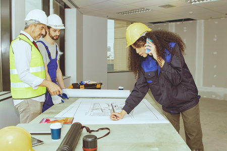 Team architects and brainstorming workers attend workshop in an office renovating and planning new construction, and female worker uses smartphone to discuss editing blueprints.の写真素材