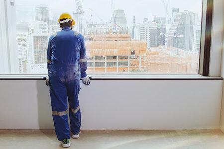Back view asian male worker working at a construction site in a tall building in the city stands looking out of a glass window and contemplates positive and inspirational accomplisの写真素材