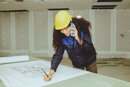 Female worker wearing helmet uses smartphone to chat with an architect foreman taking notes on the details of an office workshop office that needs renovation and planning with inteの写真素材
