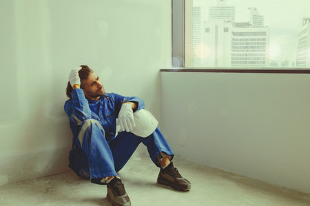 Architect asian male worker taking off his helmet confined himself in the office of an unfinished construction site sitting out of the window contemplating his thoughts feeling tirの写真素材