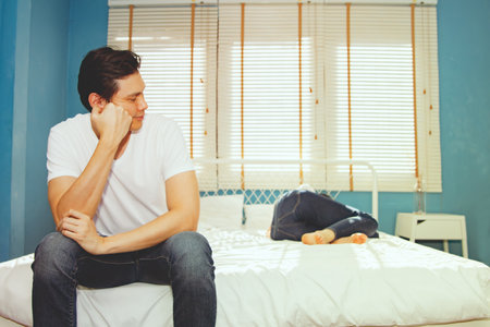 Husband who wants to have upset sex sits upset bored unhappy and uses consciousness to look at his frail wife lying sick with fever during the menstrual period, lying sick, recuperの写真素材