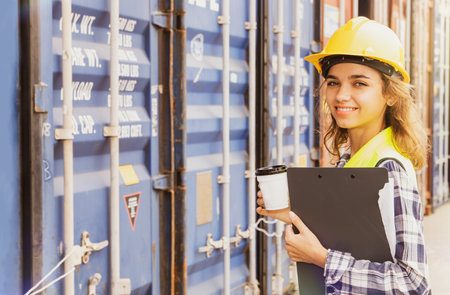 Portrait healthy caucasian young woman in uniform and helmet with folder in hand patrolling the front of the container in the working port loading yard, refreshed and alert with aの写真素材