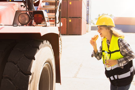 Female workers forklift drivers in the container loading yard eating hamburgers and drinking water breakfast before duty driving walking checking the safety of wheels and other parの写真素材