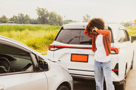 Disappointed african american woman watching her new car get crashed, rear trunk damaged standing using smartphone to call the insurance company to clear and assess the damage urgeの写真素材