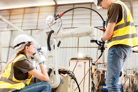 Female electrician engineer sits observing male engineer working attentively inspecting welding electrode robotic arm in auto parts factory warehouse.の写真素材