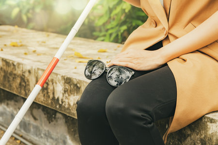 Young blind woman with vision disabilities takes off sunglasses protects eyesight and holds blind cane sitting calmly on concrete platform relaxing in park : Self care blind disablの写真素材
