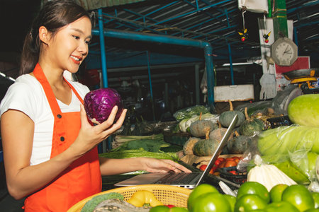 Beautiful female retailer in the market is selecting fresh vegetables for customers to order online for quick delivery : Happiness young asian female vendor sells fresh vegetablesの写真素材