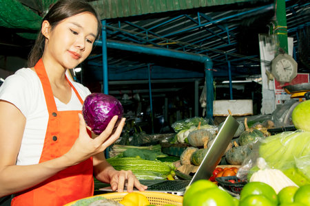Beautiful female retailer in the market is selecting fresh vegetables for customers to order online for quick delivery : Happiness young asian female vendor sells fresh vegetablesの写真素材