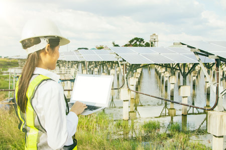 Back view asian female electrical engineer in hard hat works in the field checking solar panel installation site on suburban well, typing results on tablet online system to connectの写真素材