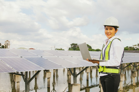 Professional asian female electrical engineer in hard hat works in the field checking solar panel installation site on suburban well typing results on tablet online system to conneの写真素材