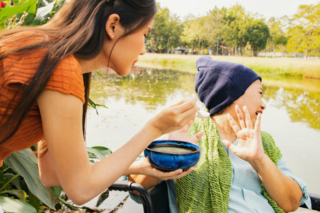 Daughter taking care health elderly mother who is sick with cancer sitting in wheelchair to eat soup to restore body and health but mother is bored of food doesn't want to eat blanの写真素材