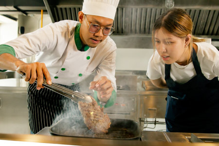 Male chefs, cooking instructors for female students in a tutorial class practice inside workshops in modern and clean kitchens cooking school teaches grilling medium rare steaks smの写真素材