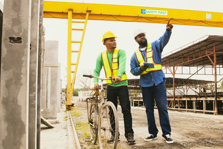 Professional African American male foreman guiding and pointing Asian male worker traveling by bicycle on factory construction site spacious precast concrete wall fast work.の写真素材