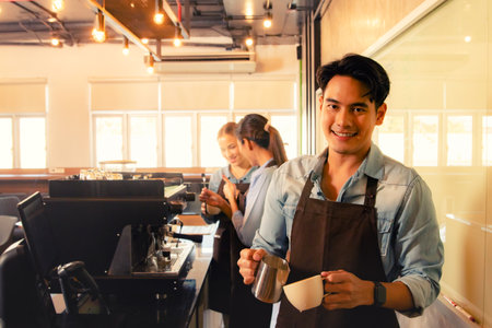 Portrait handsome young asian barista pours milk froth into latte art coffee cup decorate it beautifully appetizing mellow together with beautiful barista staff serving customers iの写真素材