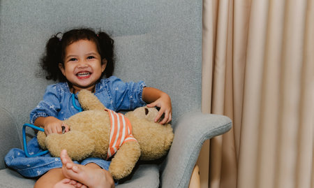 Portrait innocent little asian girl sits in a sofa chair playing with her favorite toy teddy bear and medical stethoscope in living room having humor fun without worry lovely daughの写真素材