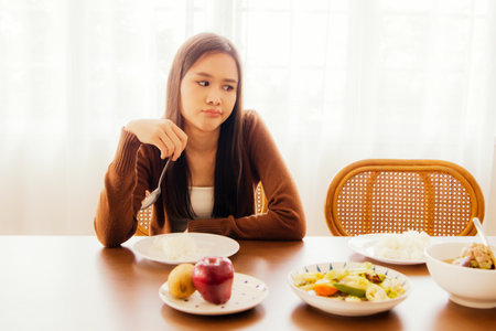 Young asian woman hungry but bored eating on table full of bland food sitting looking at food with boredom and having anorexia, not happy to eat and not having good appetite.の写真素材
