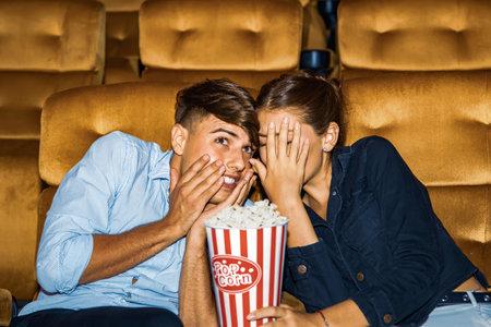 Caucasian couple holding popcorn sitting watching horror movie theater looking at a frightening scene and both them endured watching the movie with panic covering their faces withの写真素材