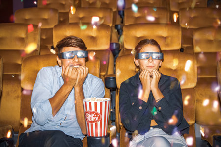 Couple puts popcorn on a chair, wears 3D glasses, sits and watches multiplex movie in an adventurous cinema looking at the scene that allows the hero to defeat the villain with botの写真素材