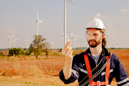Engineer male technician standing holding light bulb in wind field and looking at light bulb : Wind energy is used to generate electricity that is environmentally friendly.の写真素材