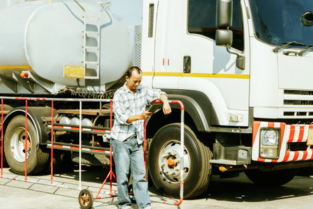 Asian male worker driving truck transport oil business industry in regional gas station checking car time on tablet parking lot to travel delivering liquid oil is on its way to desの写真素材