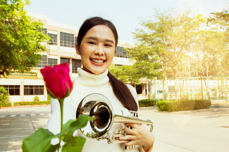 International music teacher in school : Portrait beautiful asian teacher healthy holding bright red rose and trumpet in front of school building handing flowers with friendly smileの写真素材