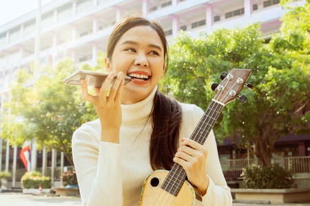 Beautiful Asian teacher sits on ukulele in front of school building preparing to teach students in...