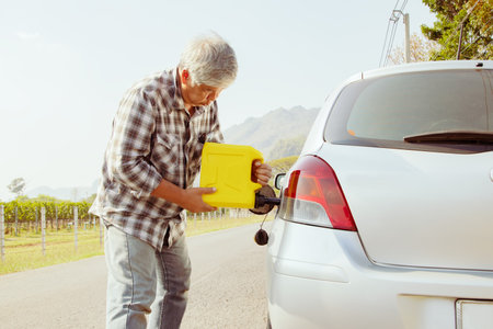Thoughtful asian senior man holding gallon of yellow fuel, spare fuel tank prepared for emergency running out of fuel while traveling on rural road without refueling station.の写真素材