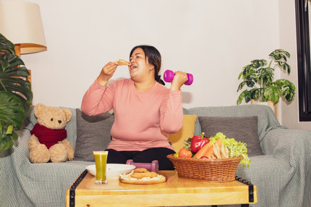 Asian fat woman in good mood eats gluttonous food to her liking prepares smoothie sits eating delicious pizza and lifts dumbbells for light exercise together in living room : Exercの写真素材
