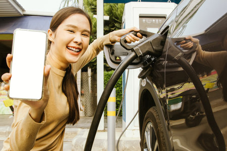 Smiling young woman in casual pose showing mobile phone with white blank screen for advertisement Introducing an application used to reserve electric vehicle charging at gas statioの写真素材