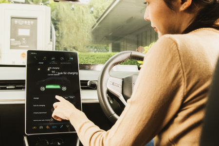 Back view young woman asian driving an EV electric car sit in the passenger compartment examining the modern touchscreen setup, observing the near full DC charging in a public fillの写真素材