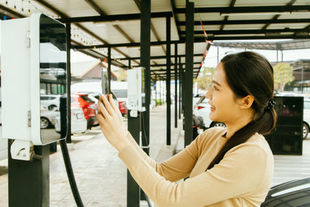 Side view Satisfied young Asian woman in a DC power station in a parking lot uses a smartphone to scan a power station bar code to charge an electric vehicle conveniently and quickの写真素材