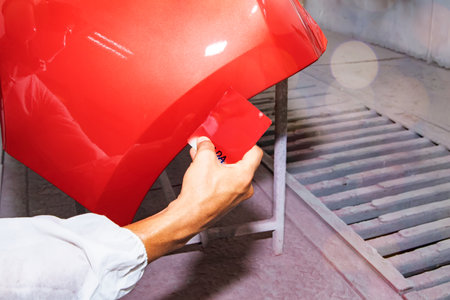 Hand of a male auto painter working car painting room inspects and compares the red paint to the sheet metal sprayed front bumper to get similar and beautiful color : Jobs in autoの写真素材