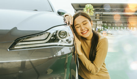 Beautiful Asian woman sitting on the side of the car in love and fascinated by EV electric car having romantic sweet dream with her new car happy and cherished face, delighted andの写真素材