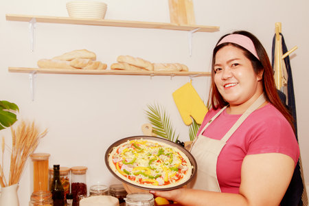 Pizza made at home : Portrait asian fat woman holding a tray of homemade pizza in the kitchen decorated with appetizing raw ham cheese being put into the oven to make the pizza cooの写真素材