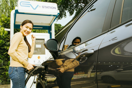 Happy smiling asian young woman raises her hands in delight as the battery of the electric car is nearly full in no time : Woman holding charger plug ready to unplug from crotch ofの写真素材