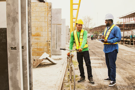 Professional African American male foreman chatting with Asian male worker traveling by bike in factory construction site, spacious precast concrete wall, work fast.の写真素材