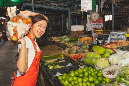 Vegetable market business : Young beautiful happy asian female seller carrying bags of tomatoes selling fresh vegetables in the market is setting up various vegetables for retail sの写真素材