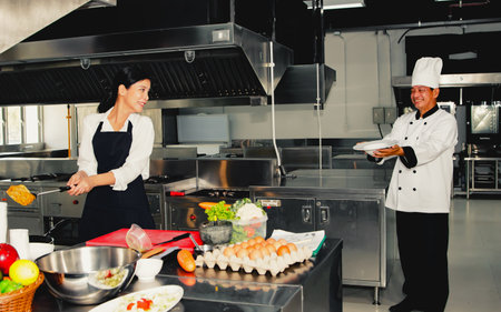 Happiness in the kitchen : Senior male chef stands in a good mood to receive the fried eggs cooked by the beautiful young female student chef in the kitchen to toss and receive theの写真素材