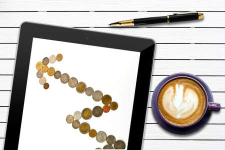 arrow from coins showing investment rise on tablet,pen and coffee cup on wood background